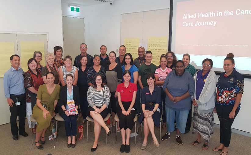 26 people standing together smiling in a presentation room.