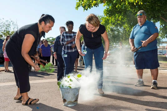 Two women carrying out a smoking ceremony, with many First Nations people observing them. A man stands beside them holding clapsticks.