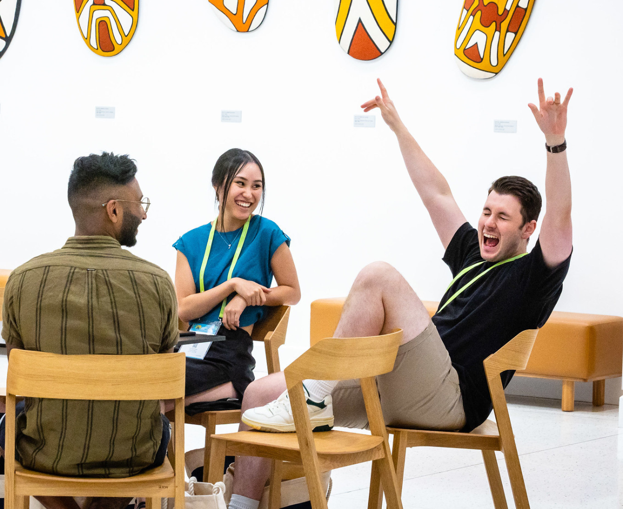 Three people sitting around a table, looking excited. The right-most man has his hands in the air and appears to be shouting with joy.